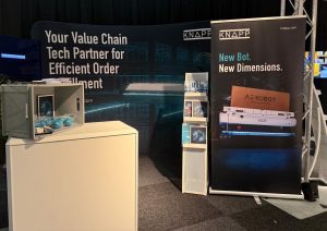 Beursstand voor KNAPP met banners over efficiënte orderafhandeling en een nieuwe robot genaamd AEROBOT, met een vitrine met blauwe ballen en brochures op een witte balie en stand.