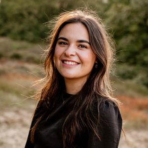 Een jonge vrouw met lang bruin haar lacht naar buiten en draagt een zwart topje. De achtergrond is wazig en laat groen en zonlicht zien.