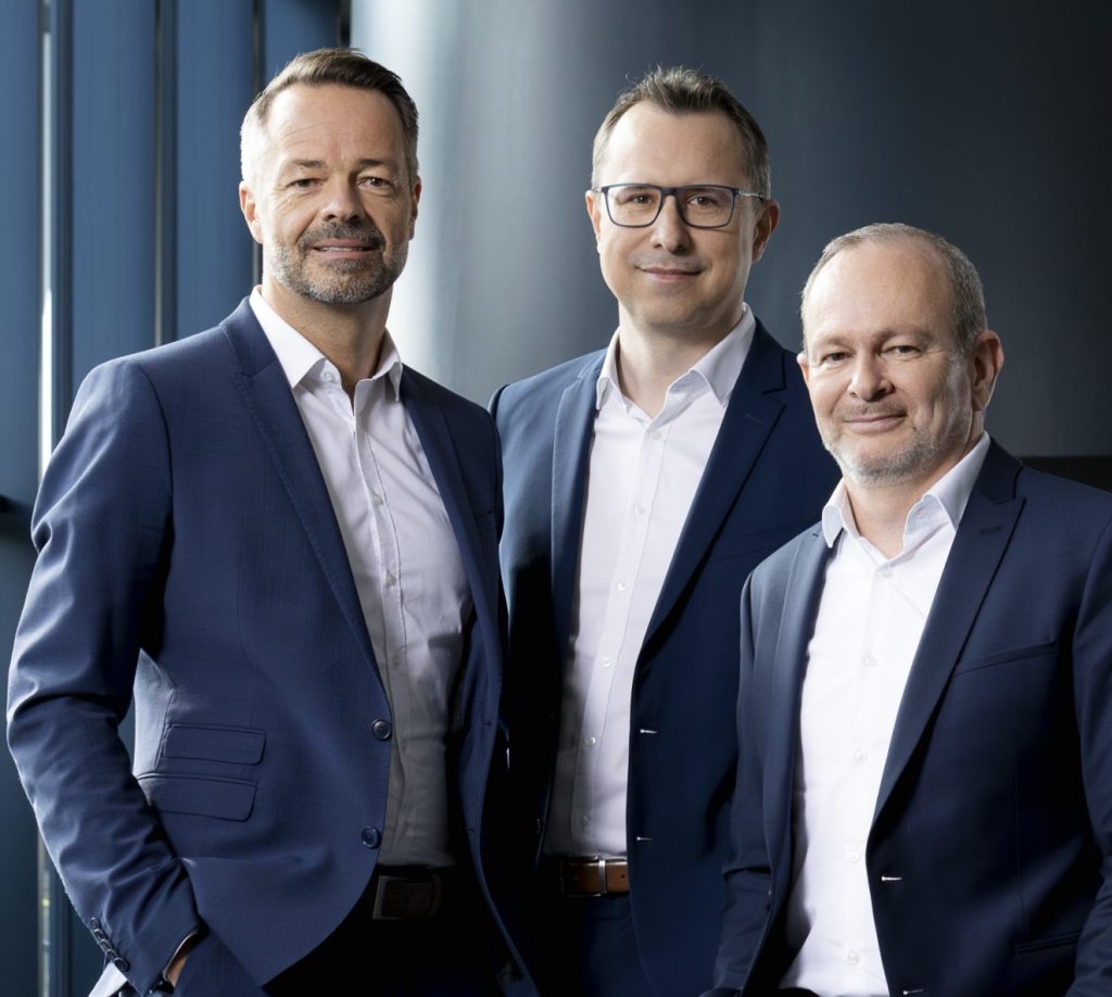 Drie mannen in zakelijke pakken en witte overhemden poseren samen, glimlachen lichtjes, in een moderne kantooromgeving met blauwe en grijze tinten op de achtergrond.