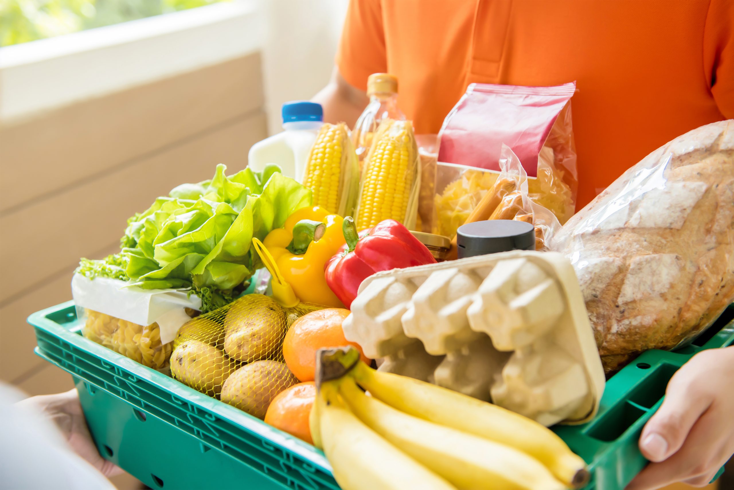 Een persoon in een oranje shirt houdt een groene krat vast die gevuld is met boodschappen, zoals bananen, brood, eieren, melk, verse groenten, pasta en drank in flessen. Een andere hand reikt naar de krat.