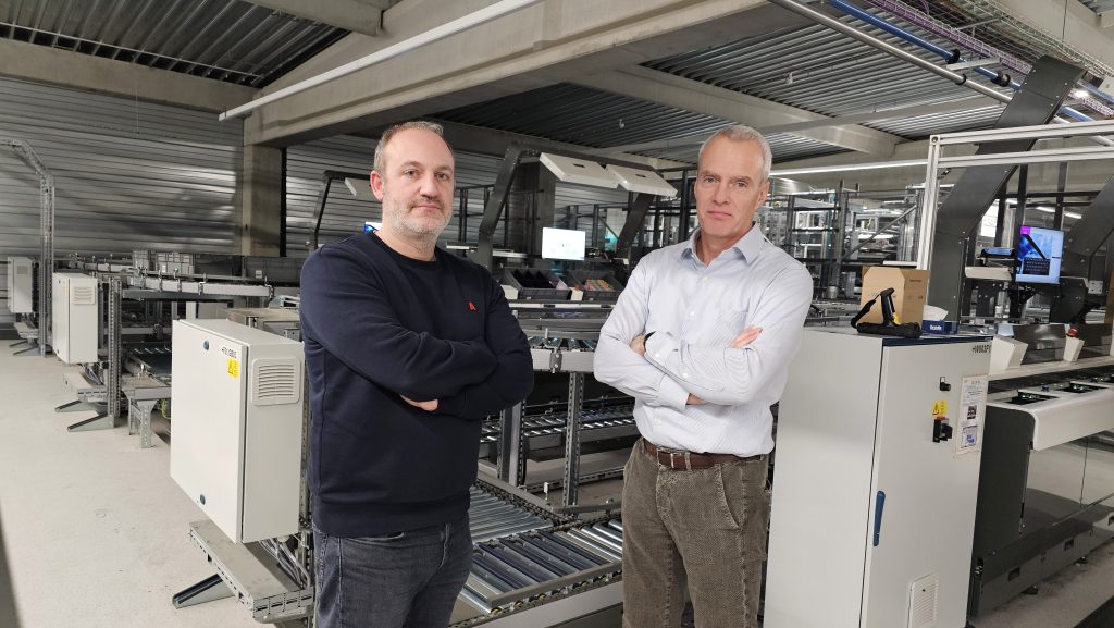 Twee mannen staan met gekruiste armen in een moderne industriële omgeving, omringd door geautomatiseerde machines en lopende banden. Beiden kijken zelfverzekerd naar de camera, wat een professionele en collaboratieve omgeving weerspiegelt.