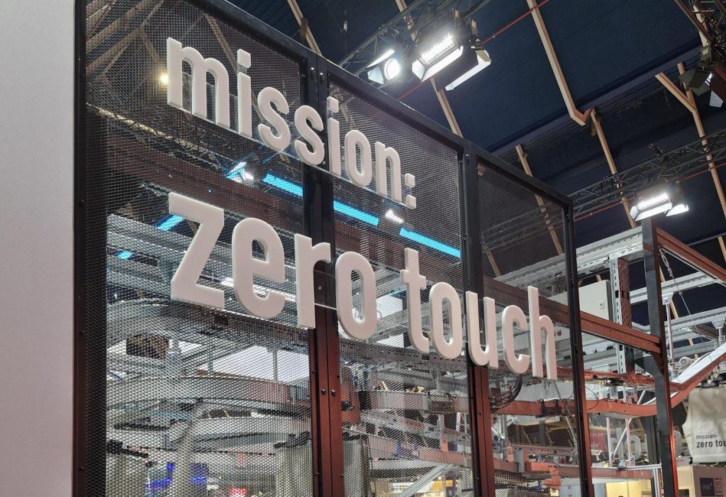 Grote witte letters op een scherm van metaalgaas spellen "missie: zero touch" op een overdekte industriële of technologische expo, met machines en plafondverlichting zichtbaar op de achtergrond.