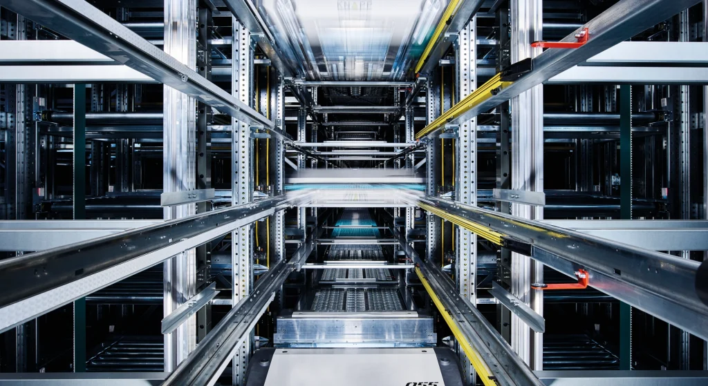 Zicht in een geautomatiseerd magazijnsysteem met metalen rekken en rails, waarbij een robot of shuttle over de rails beweegt, omgeven door reflecterende metalen balken en industriële verlichting.