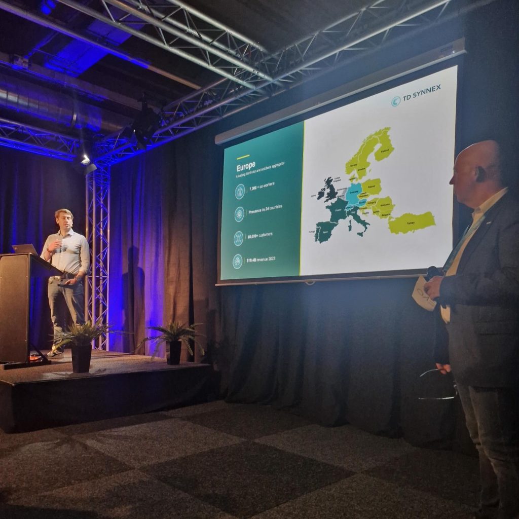 Twee mannen presenteren op het podium voor een groot scherm met een kaart van Europa met gemarkeerde landen en het TD SYNNEX logo. De setting lijkt een conferentie of bedrijfsevenement met blauwe podiumverlichting.