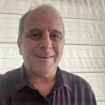 A middle-aged man with short gray hair smiles at the camera, wearing a dark plaid shirt. He is standing in front of a white, horizontal-panel background.