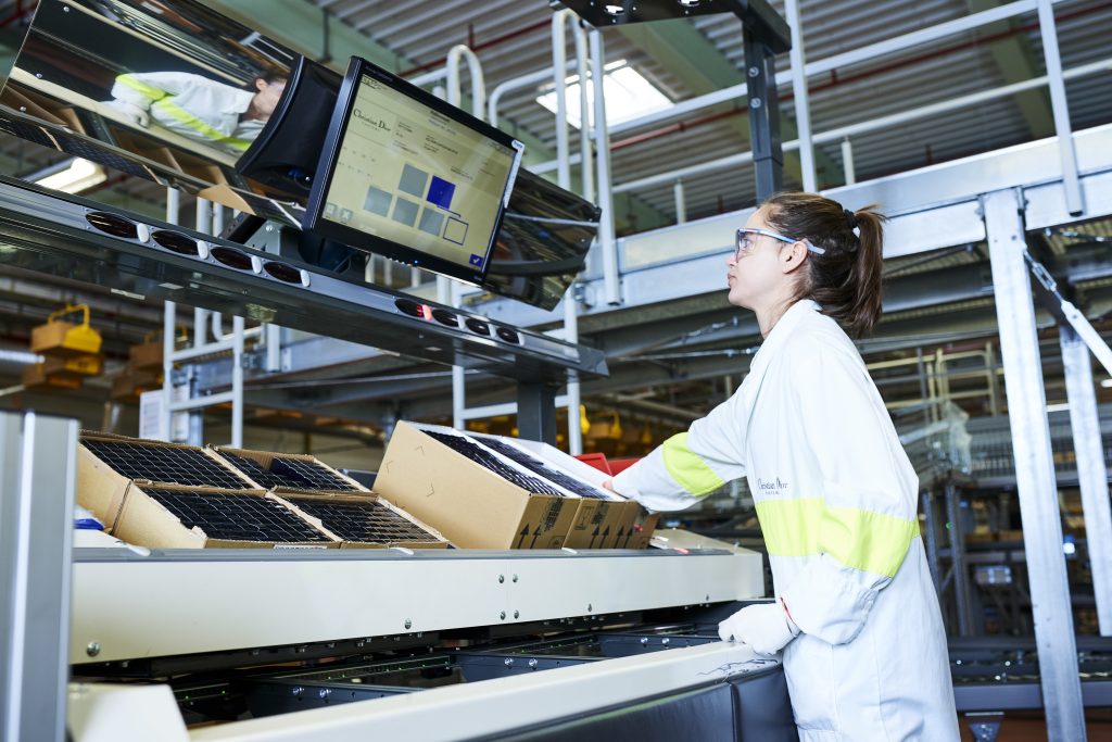 Een vrouw met een veiligheidsbril op en een labjas aan bedient een transportsysteem en sorteert pakketten in een magazijn terwijl ze naar een computerscherm kijkt waarop sorteerinformatie wordt weergegeven.