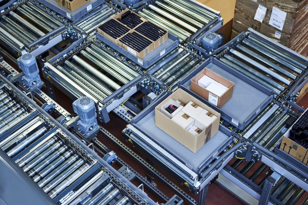 Dozen met verschillende producten rijden op een transportband in een magazijn of distributiecentrum, omringd door metalen rollen en machines.