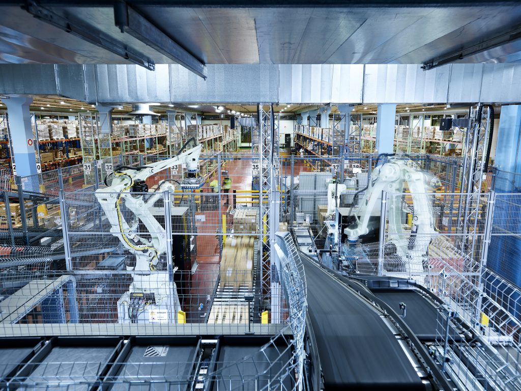 Een groot, modern magazijn met industriële robots en transportbanden die pakketten sorteren. Op de achtergrond zijn rekken met dozen zichtbaar en het gebied is omheind met veiligheidshekken. De scène is helder verlicht en georganiseerd.