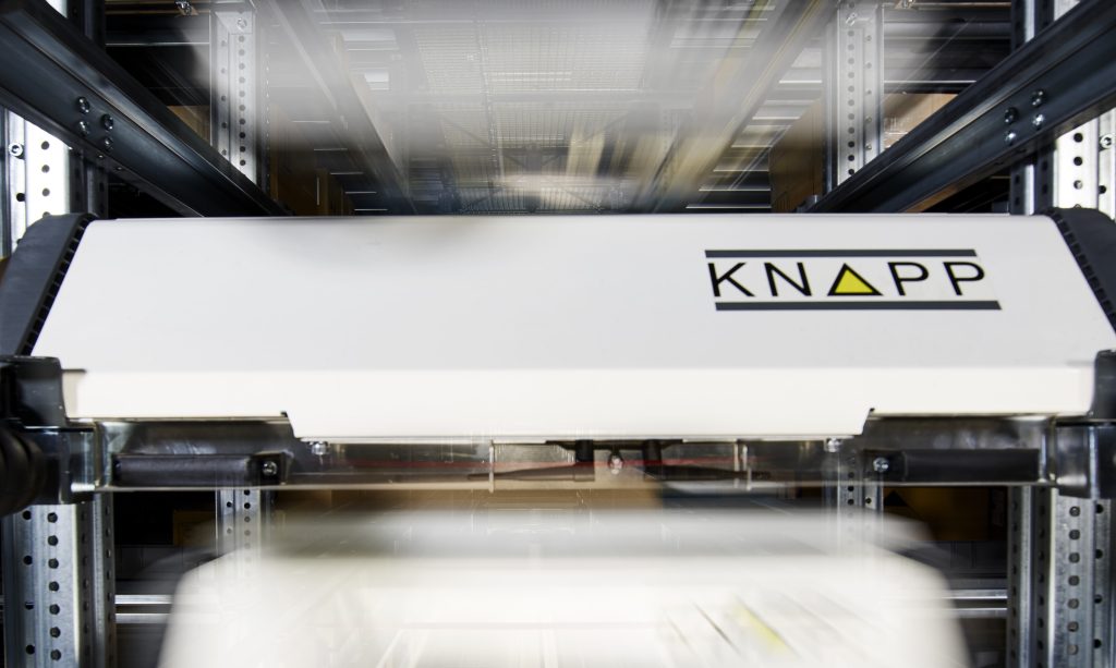 Een witte geautomatiseerde magazijnrobot met het label "KNAPP" beweegt snel over metalen rupsbanden, waarbij de onscherpe beweging de snelheid benadrukt in een industriële opslagomgeving.