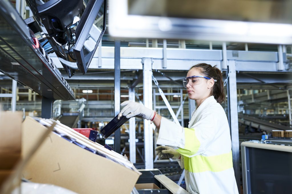 Een vrouw draagt een veiligheidsbril, handschoenen en een veiligheidsvest en werkt aan een transportband in een industriële faciliteit, waar ze verpakkingen verwerkt in de buurt van een kartonnen doos en computerschermen.
