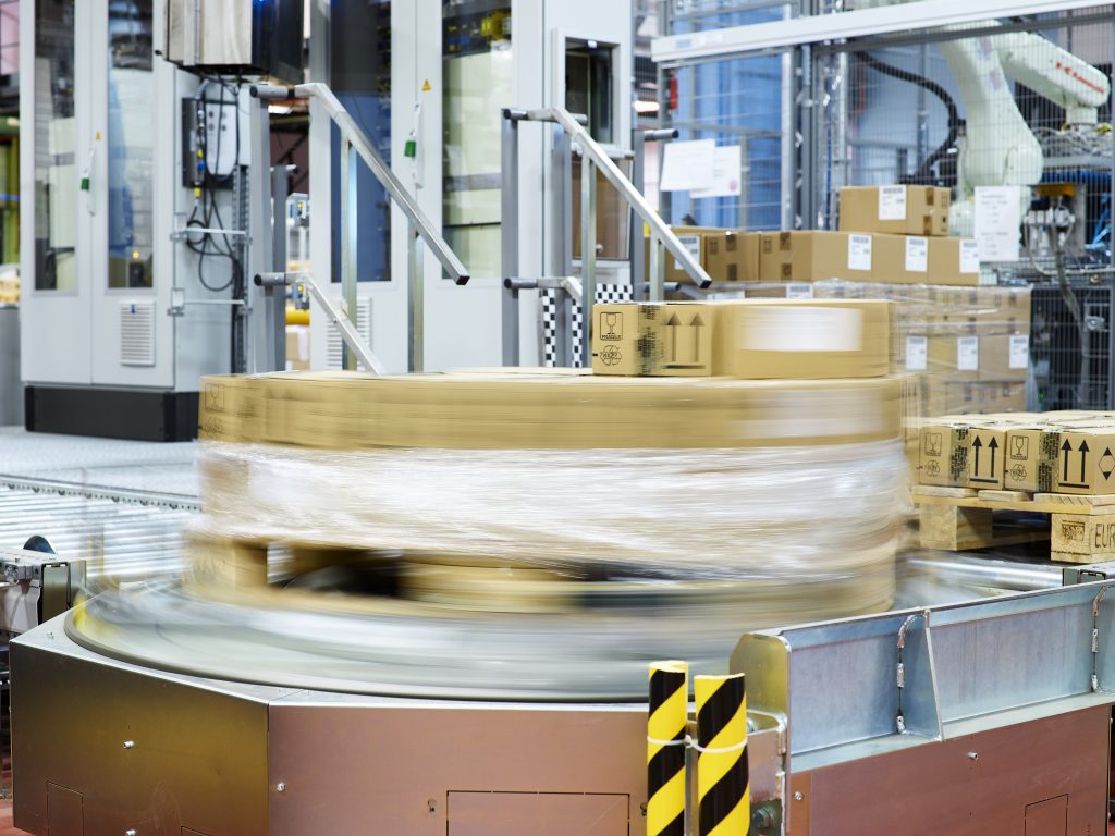 Een pallet met kartonnen dozen draait snel rond op een geautomatiseerde machine in een fabriek en wordt verpakt in doorzichtig plastic voor verzending. Op de achtergrond zijn industriële apparatuur en gestapelde dozen te zien.