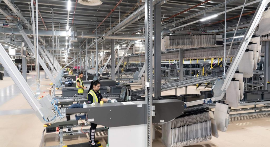 Case study the very group - visual 1 Werknemers in veiligheidsvesten bedienen machines en computers in een groot, modern magazijn of distributiecentrum met rijen metalen rekken en geautomatiseerde systemen.
