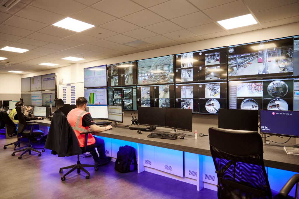 Case study the very group - visual 2 Een beveiligingscontrolekamer met meerdere mensen die meerdere grote schermen in de gaten houden waarop beelden en gegevens van beveiligingscamera's worden getoond. Eén persoon in een roze vest zit aan een bureau met computers, tegenover de videomuur.