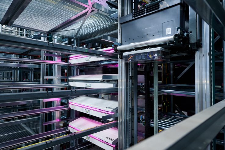 Case study the very group - visual 4 Een modern geautomatiseerd magazijnsysteem met metalen rekken en robotplateaus die worden verlicht door paarse LED-lampen, die een geavanceerd opslag- en zoekmechanisme weergeven.