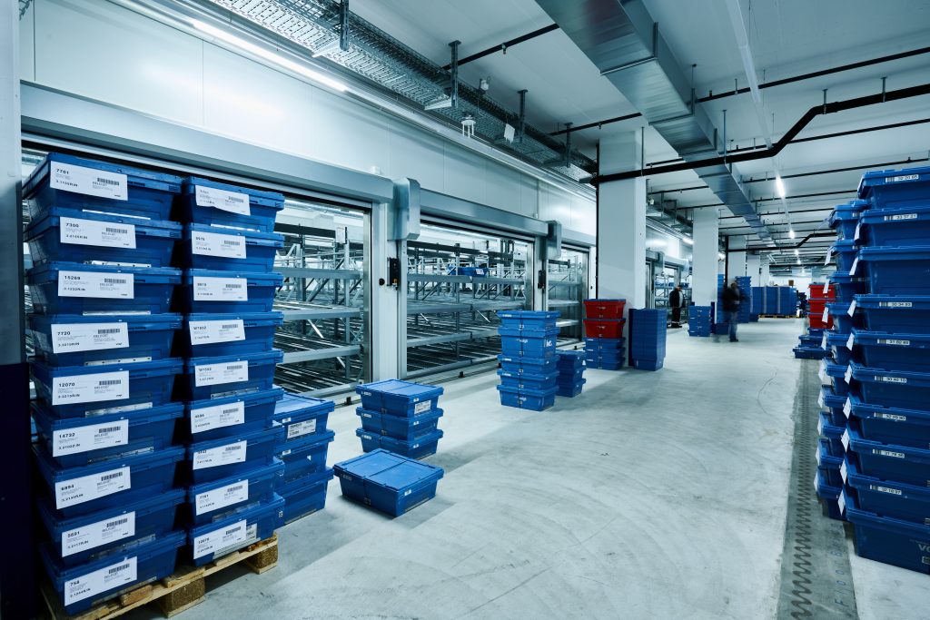 Case study VOIGT - visual 8 Magazijninterieur met stapels blauwe plastic bakken en kratten, sommige gelabeld met witte labels, gerangschikt op pallets en de vloer. Metalen rekken zijn zichtbaar op de achtergrond onder heldere verlichting.