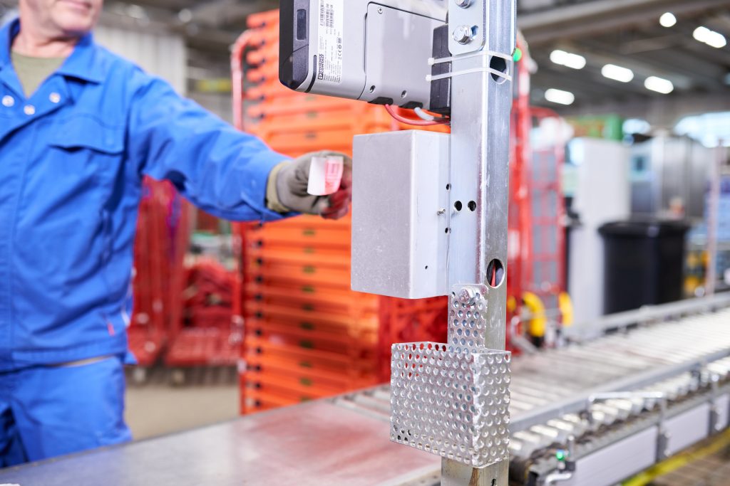 BN Lichtbilder Een arbeider in een blauw uniform gebruikt een sleutelkaart om toegang te krijgen tot machines op een industriële productielijn, met apparatuur en rode rekken op de achtergrond.