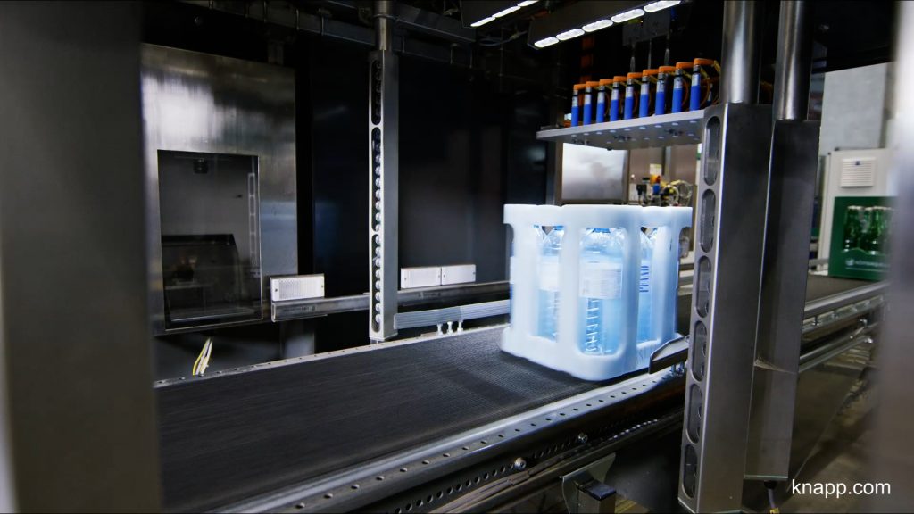Cleanaway visual 2 Een verpakking van gebotteld water verpakt in plastic beweegt over een geautomatiseerde transportband in een moderne industriële faciliteit. Metalen constructies en machines zijn zichtbaar op de achtergrond.