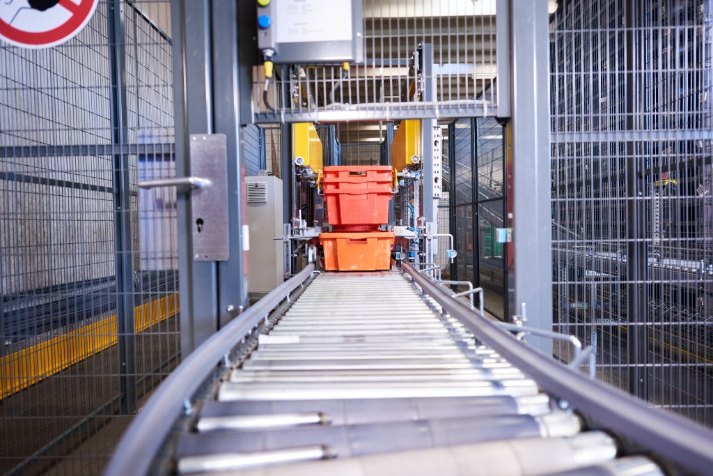Cleanaway visual 1 nl Een rode plastic container beweegt over een metalen transportband in een omheinde industriële faciliteit, met machines en veiligheidshekken zichtbaar op de achtergrond.