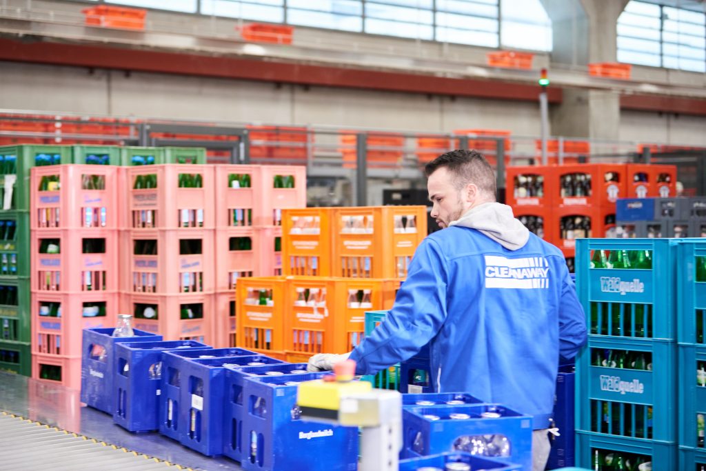 Cleanaway visual 2 nl Een man in een blauwe jas werkt in een magazijn en organiseert kleurrijke kratten met glazen flessen. De kratten staan in rijen op industriële rekken.