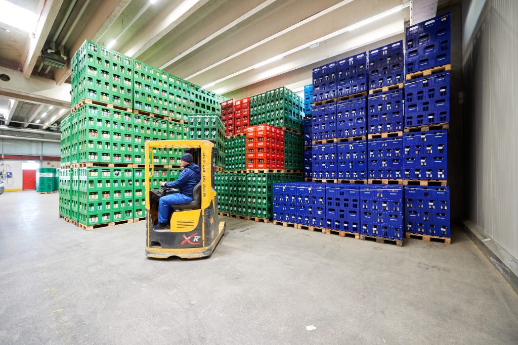 Cleanaway visual 3 nl Een persoon bestuurt een gele vorkheftruck in een magazijn omringd door grote stapels groene, blauwe en rode plastic kratten op pallets. De helder verlichte ruimte heeft betonnen vloeren en zichtbare plafondbalken.