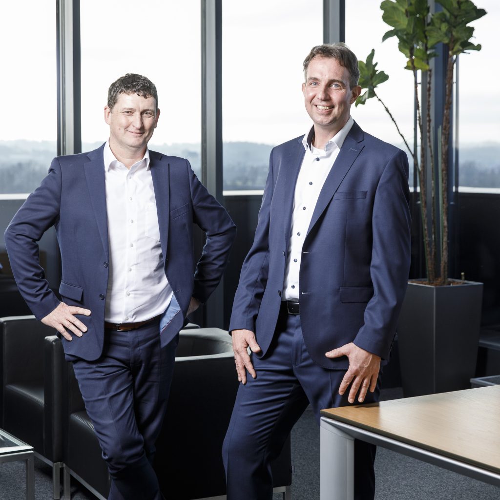 Twee mannen in blauwe pakken staan glimlachend in een modern kantoor met grote ramen, zwarte stoelen en een hoge groene plant op de achtergrond.