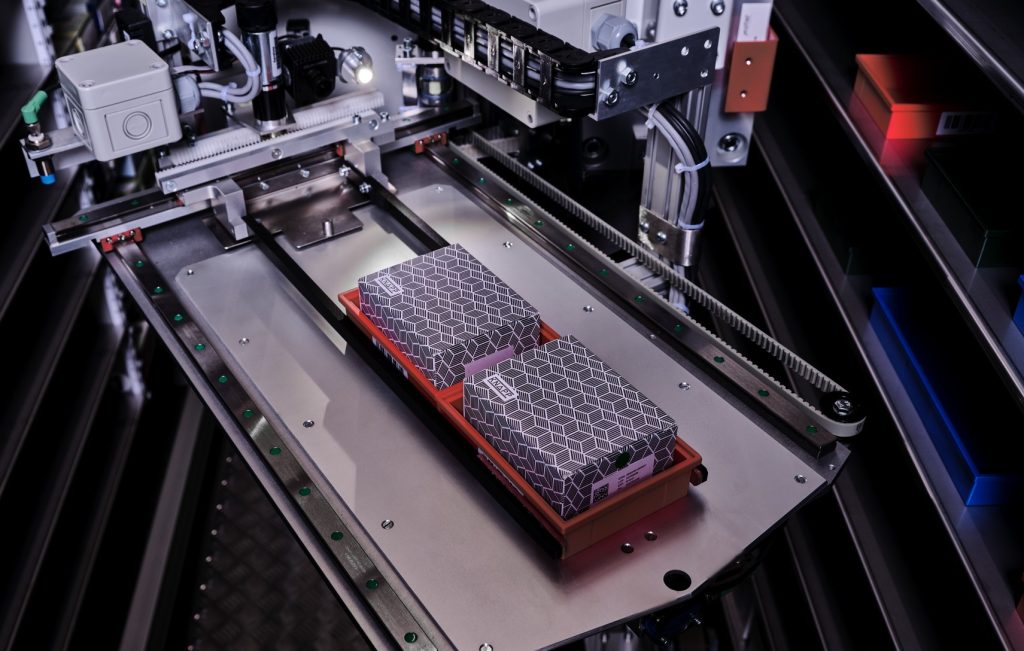 Een close-up van een industriële machine die twee voorwerpen met hexagon-patroon op een dienblad hanteert in een high-tech fabriek van KNAPP-sectoren.