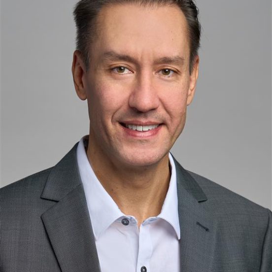 Een man met kort bruin haar draagt een grijs colbert en een wit overhemd en lacht voor een effen grijze achtergrond.