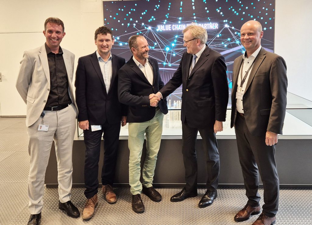 Vijf mannen in zakelijke kleding staan binnen bij elkaar. Twee van hen schudden elkaar de hand en glimlachen. Een digitaal scherm achter hen toont de woorden "Value Chain Partner" met een afbeelding van een verbonden netwerk.