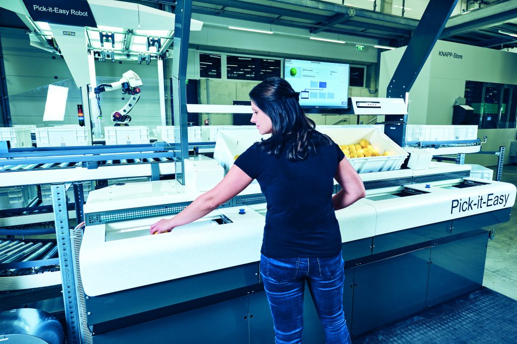 Een vrouw bedient een automatische sorteermachine met het label "Pick-it-Easy" in een moderne industriële faciliteit van KNAPP-sectoren, met op de achtergrond bakken met artikelen en een robotarm.