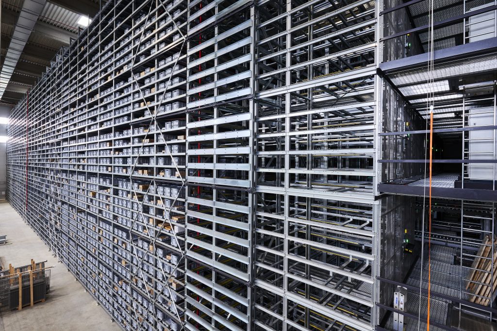 BN Lichtbilder Een groot geautomatiseerd stellingsysteem met meerdere niveaus en lege metalen rekken strekt zich hoog en lang uit in een ruim industrieel gebouw. Op de betonnen vloer zijn steigers en apparatuur zichtbaar.