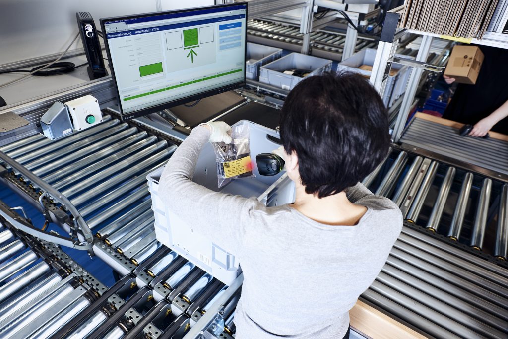 BN Lichtbilder Een persoon met kort donker haar gebruikt een barcodescanner om items in een grijze bak op een transportband te scannen, terwijl een computerscherm de inventarisgegevens voor hem/haar weergeeft.