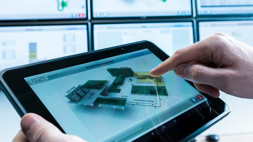 Een persoon gebruikt een touchscreen-tablet met een overzichtsdiagram van een KNAPP-sectorenmagazijn en wijst naar een sectie met het label "Visual Case Picking", met computermonitoren zichtbaar op de achtergrond, wat innovatie in de sector weerspiegelt.