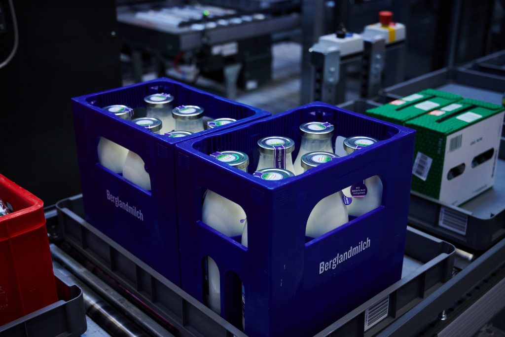 Blauwe kratten met het label "Berglandmilch" met daarin glazen melkflessen worden op een transportband geplaatst in een industriële omgeving, wat doet denken aan de automatisering die te zien is in de Case Study SPAR Ebergassing met KNAPP Benelux Automatisering.