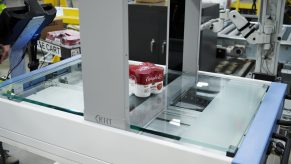 spar - visual 2 Een pak Campbell's soep staat op een glazen platform in een machine, waarschijnlijk voor scannen of kwaliteitscontrole, in een fabriek of industriële omgeving met apparatuur op de achtergrond.