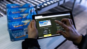 spar - visual 3 Een persoon gebruikt een tablet om productinformatie te bekijken voor een pak ingeblikte goederen met het label "Roasted Oyster", dat op een lopende band op de achtergrond staat.