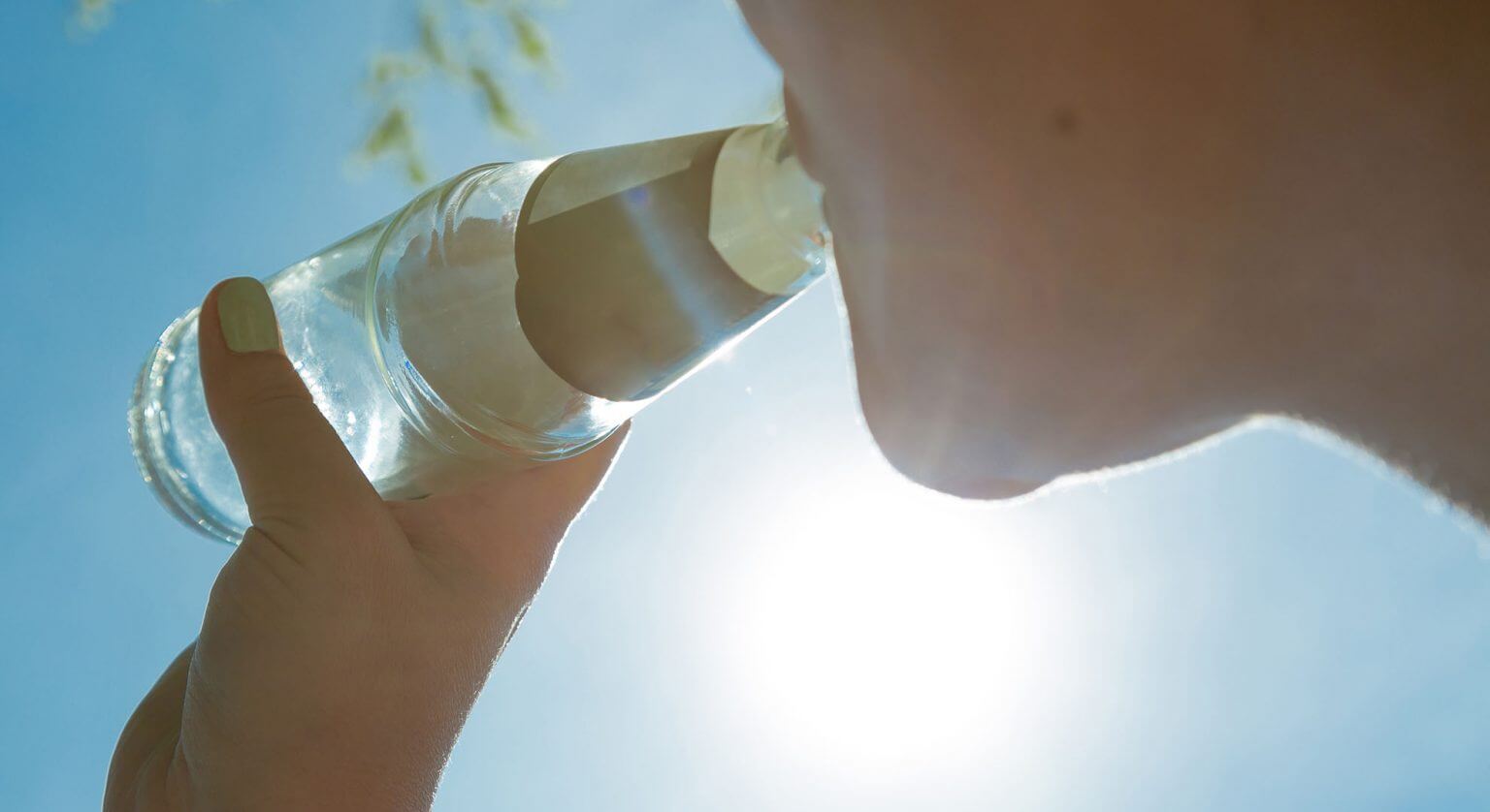 Een persoon drinkt buiten uit een glazen fles met zonlicht dat fel schijnt op de achtergrond. Hun hand houdt de fles vast en de blauwe lucht is erboven zichtbaar.